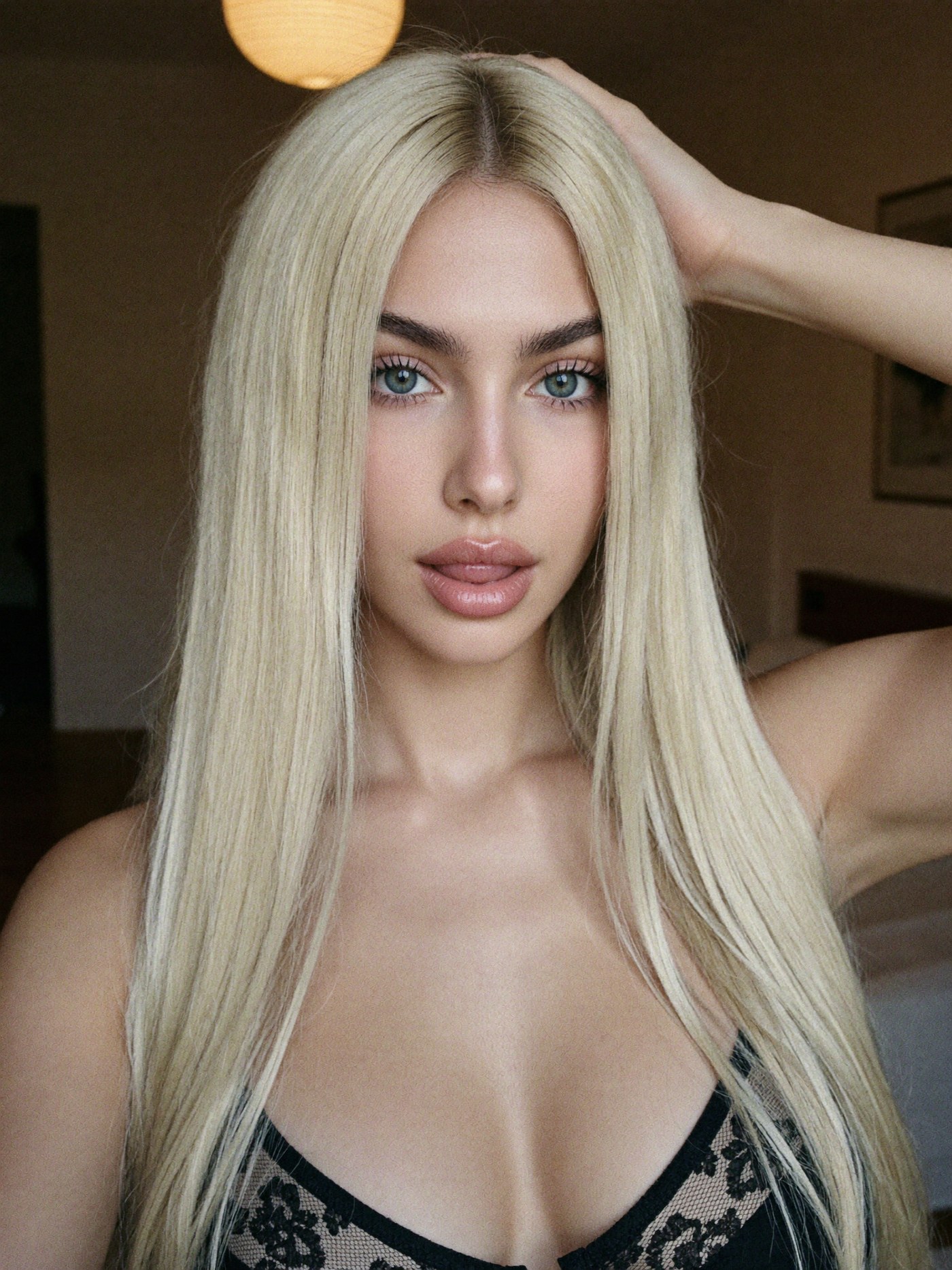 a close-up selfie from the chest up in a black lace bra, lips softly parted and eyes on the lens, one hand resting on top of her long straight platinum-blonde hair, a round paper lamp glowing above her, warm lamp light