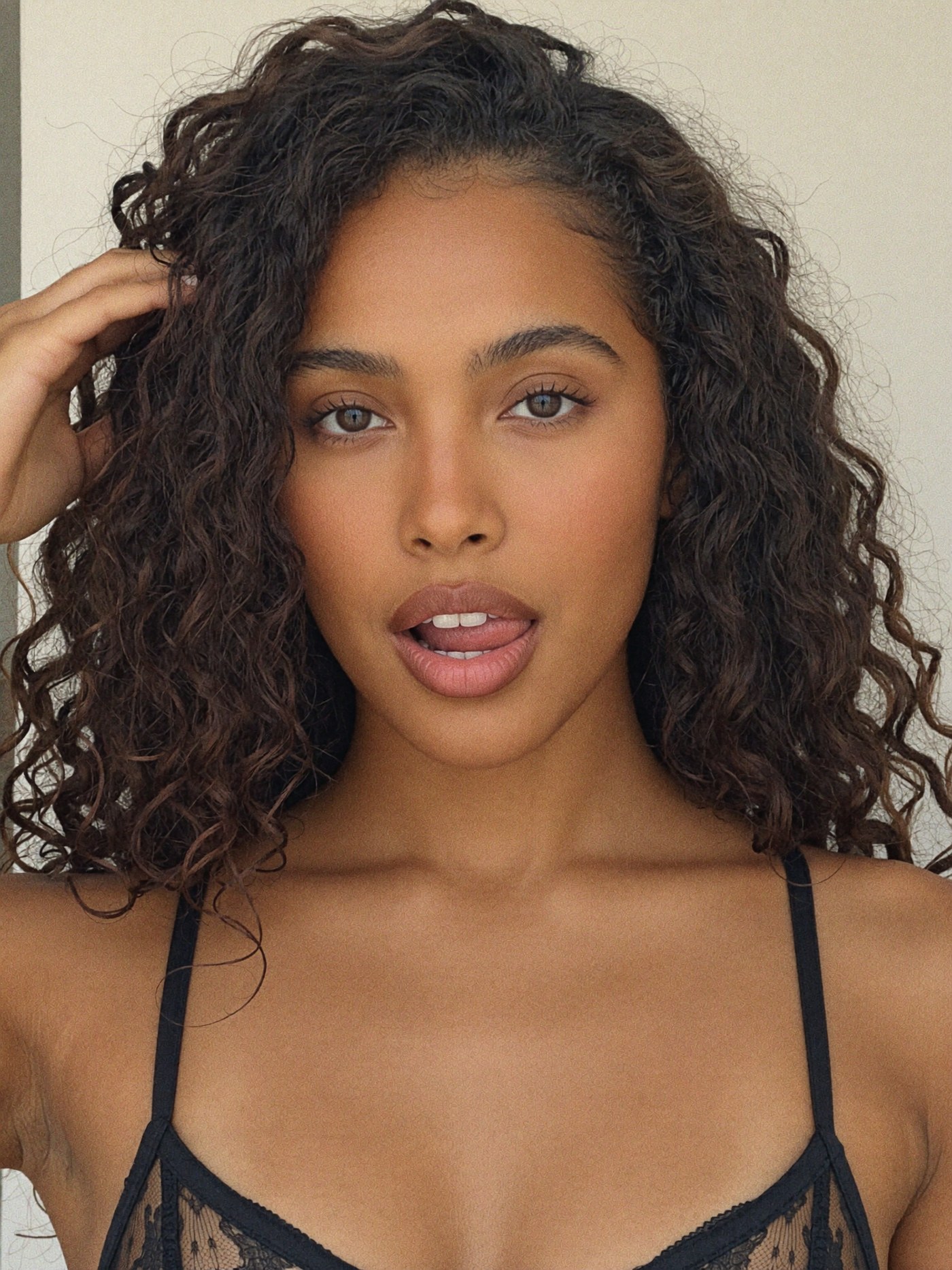 a close-up selfie from the chest up in a black lace bra, lips parted with the tip of her tongue showing, one hand pushing back her long curly dark-brown hair at the side, plain indoor wall behind her, soft daylight