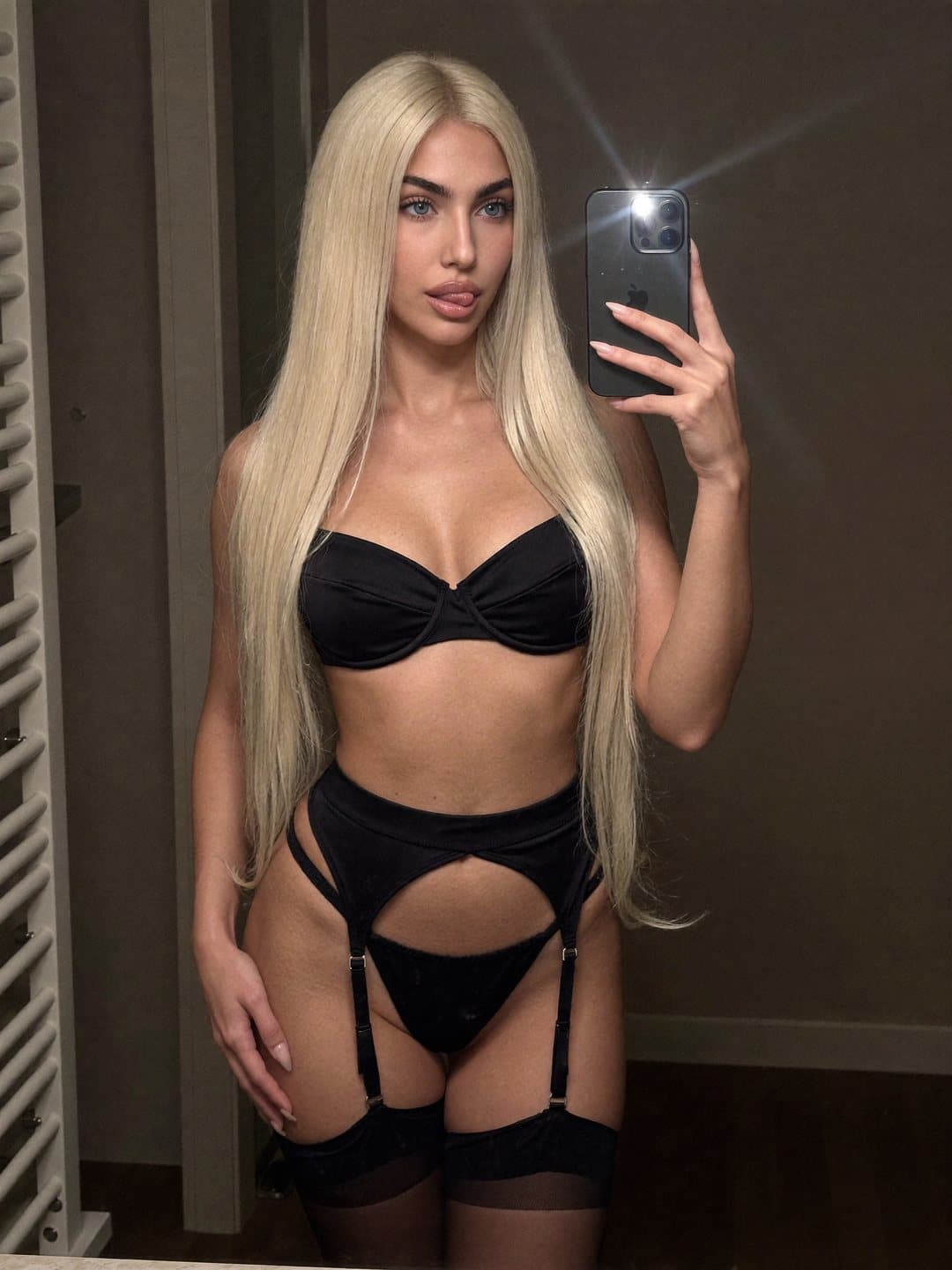 Mirror selfie of Alisa, a blonde Russian woman, with flash in a black satin balconette bra, black garter belt, black briefs and sheer black stockings, phone raised beside her face, one hand at her thigh, lips in a pout.