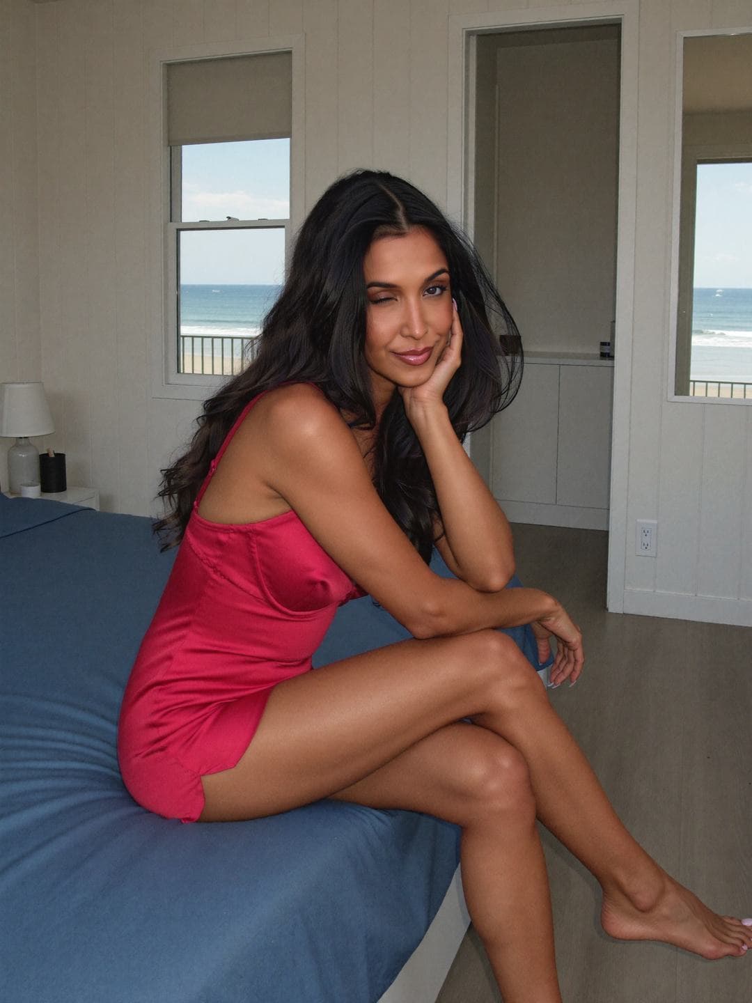 Ananya, a dark-haired Indian woman, in a short red satin slip sitting on the edge of a bed with a blue cover, legs crossed and chin on her hand, winking, a window showing the beach and sea behind her.