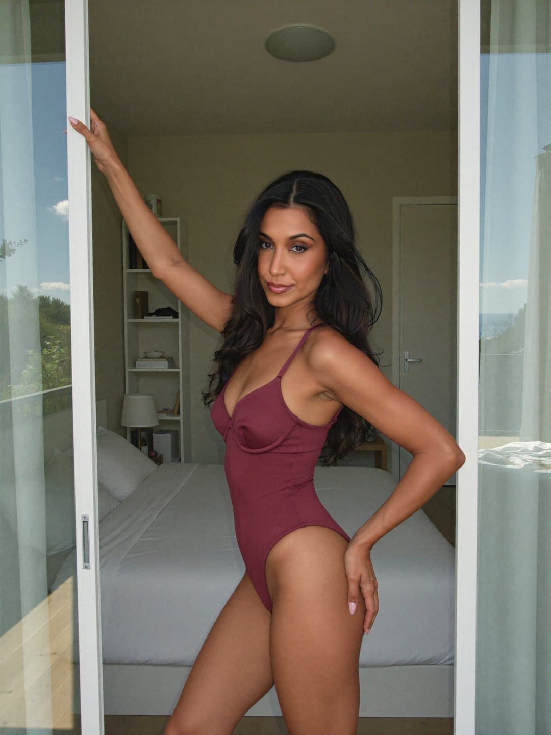 Ananya, a dark-haired Indian woman, in a wine-coloured bodysuit, stands side-on in an open glass doorway gripping the white frame overhead, her free hand on her hip, looking across at the camera with a neat bed and bookshelf inside.