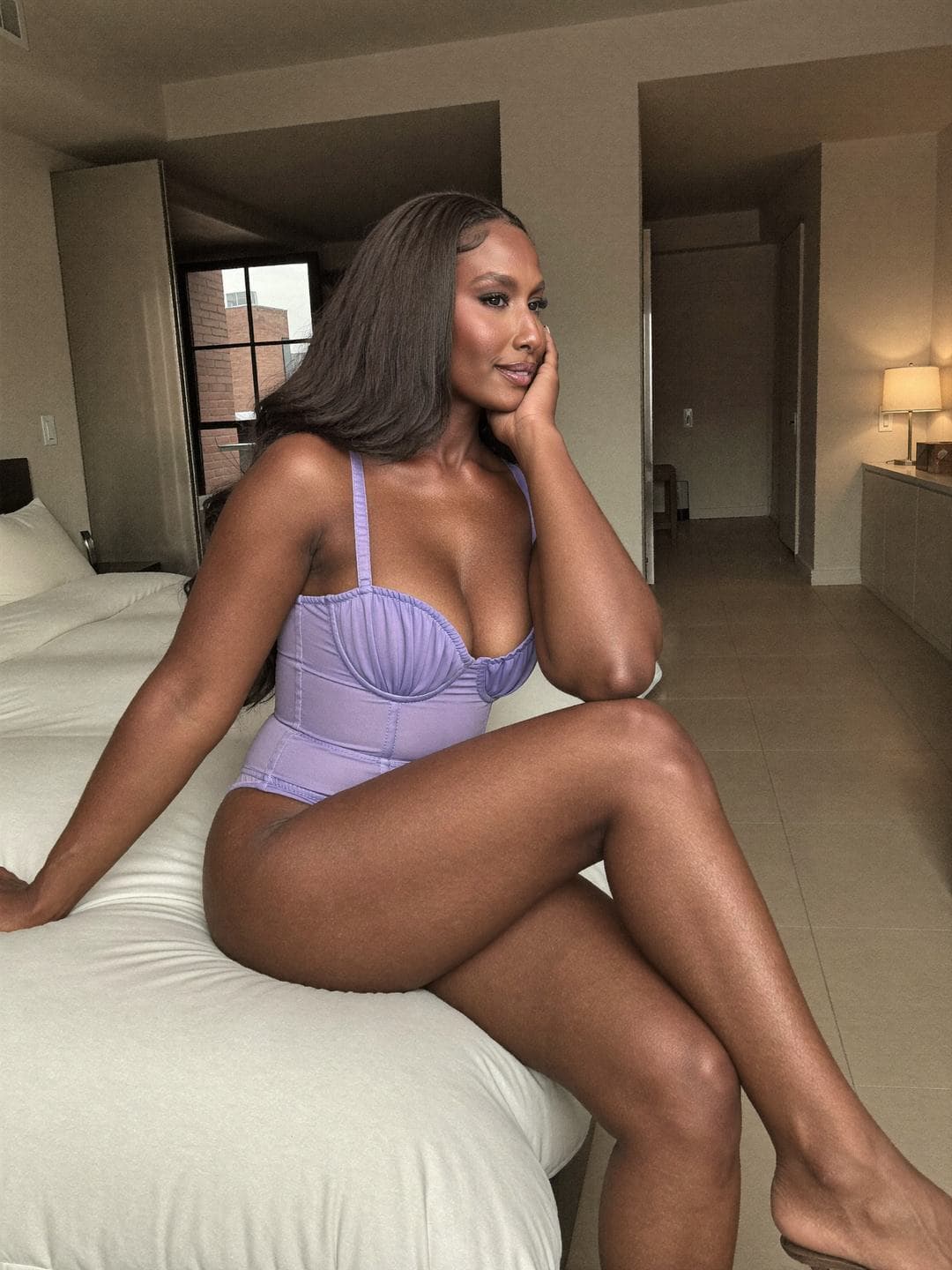 Brielle, a dark-haired African American woman, sits on a white bed in a lilac corset-style bodysuit with gathered cups, legs crossed, one elbow on her knee and her cheek in her hand, a brick window and hallway behind her, looking away dreamily.