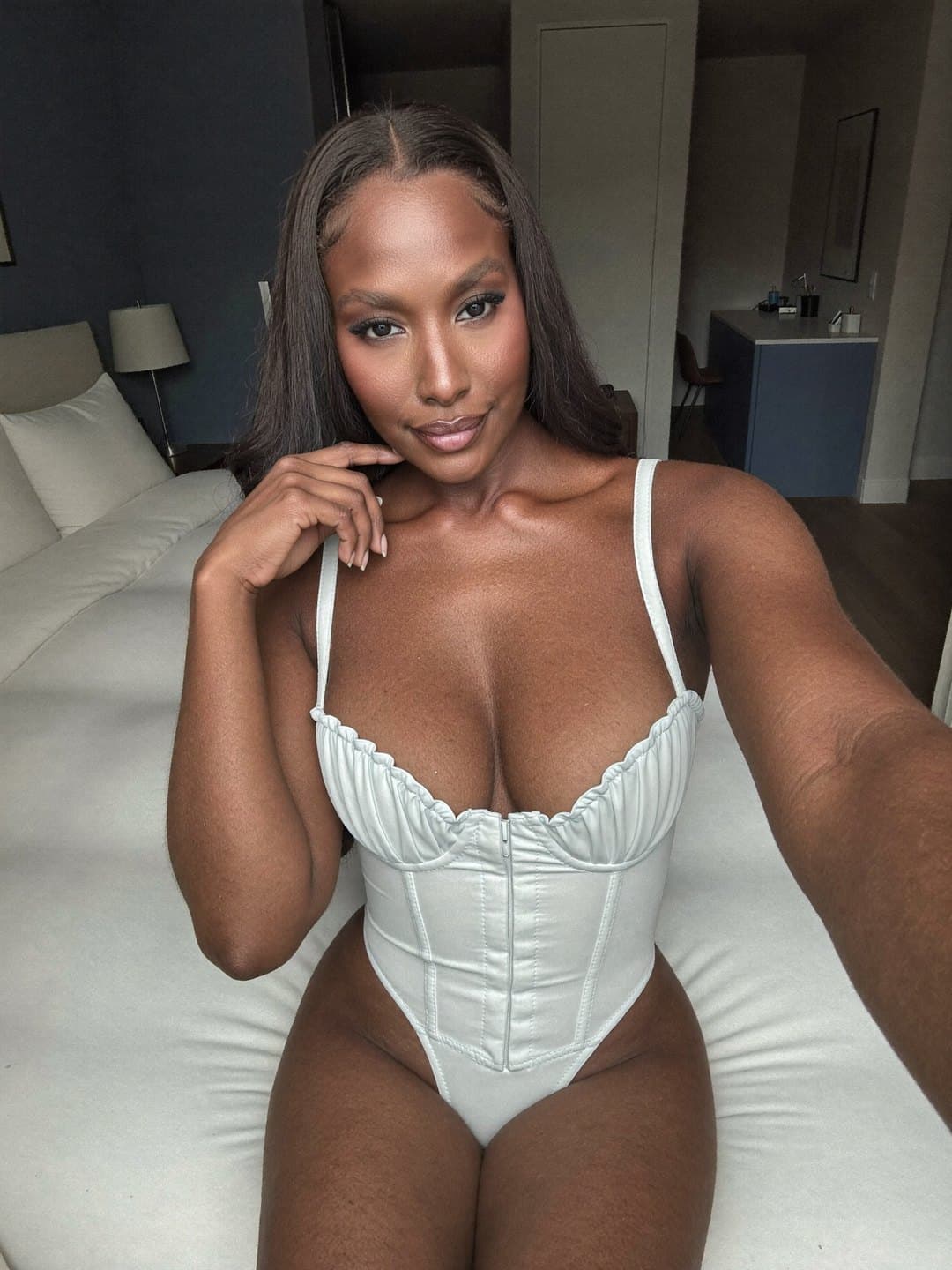 Brielle, a dark-haired African American woman, in an ivory corset bodysuit with ruched cups and a front zip takes an arm's-length selfie seated on a white bed, one hand at her chin, smiling into the lens, a navy wall and blue cabinet behind.
