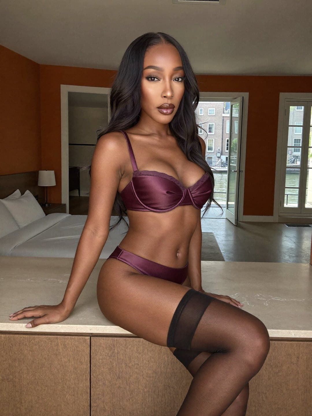 Mulberry satin bra trimmed in lace, briefs to match and sheer black stockings; Fabienne, a dark-haired Haitian woman, sits with crossed legs on a stone-topped oak counter, both hands spread wide, as open doors reveal the canal.