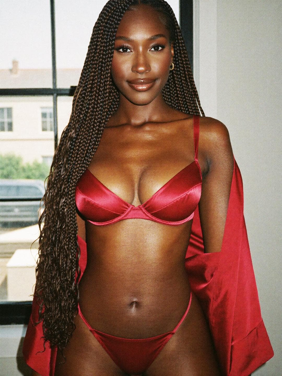 Imani, a brunette African American woman, in red satin lingerie and a red robe by a sunlit window