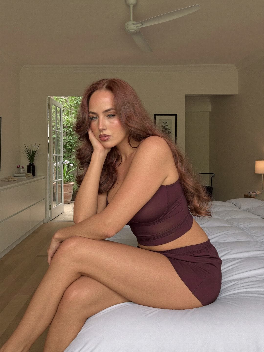 Jacinta, a redheaded Australian woman, sits on the edge of a white bed in a plum mesh-panel camisole and plum shorts, legs crossed, cheek resting on her hand, sultry pout, a white dresser and open French door to a green courtyard behind.