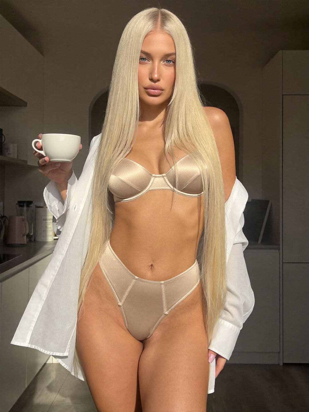 Sofiia, a blonde Ukrainian woman, in champagne satin lingerie and an open white shirt holding a cup in a sunny kitchen