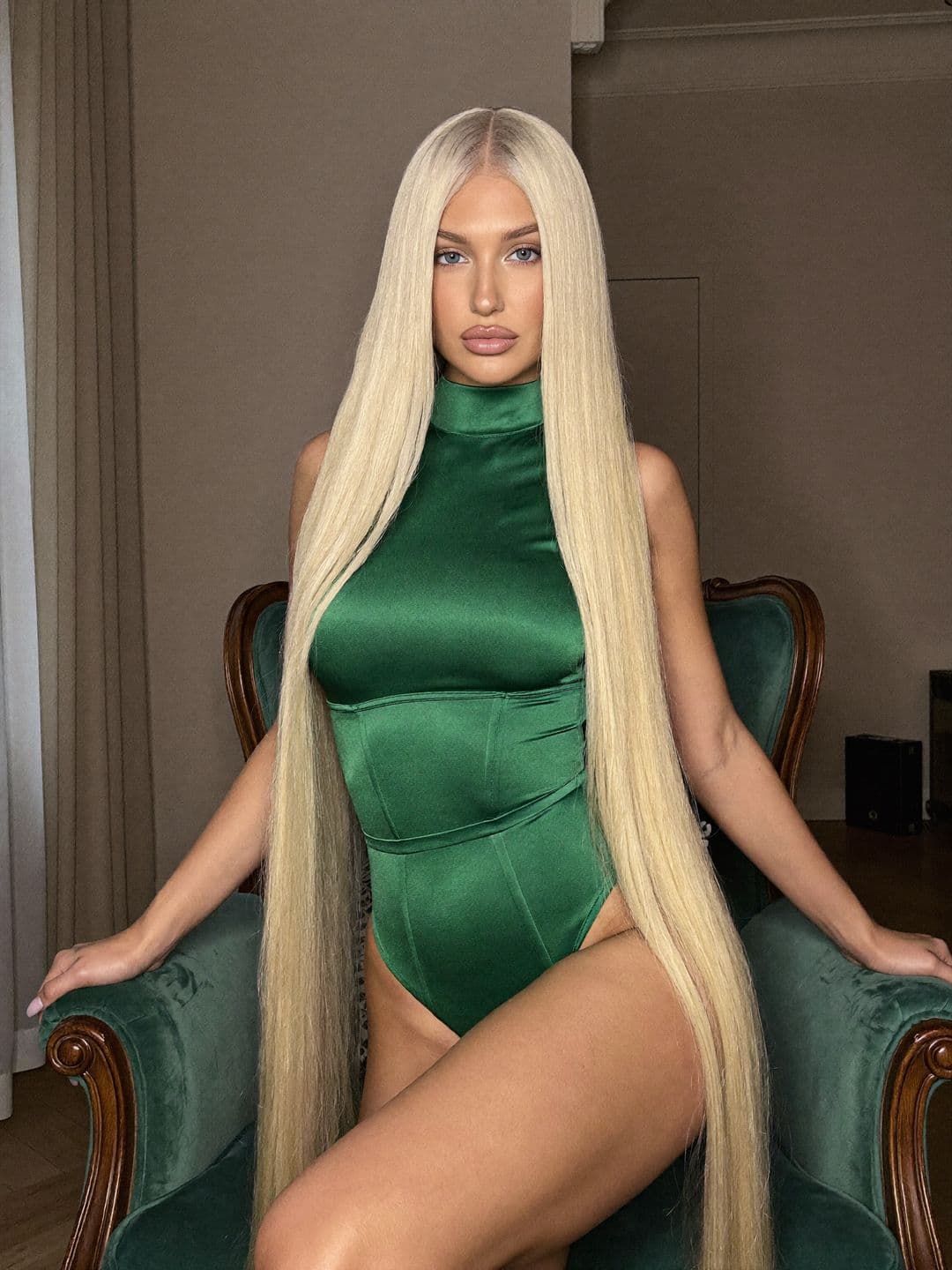 Sofiia, a blonde Ukrainian woman, in a high-neck emerald satin bodysuit sitting upright in a green velvet armchair