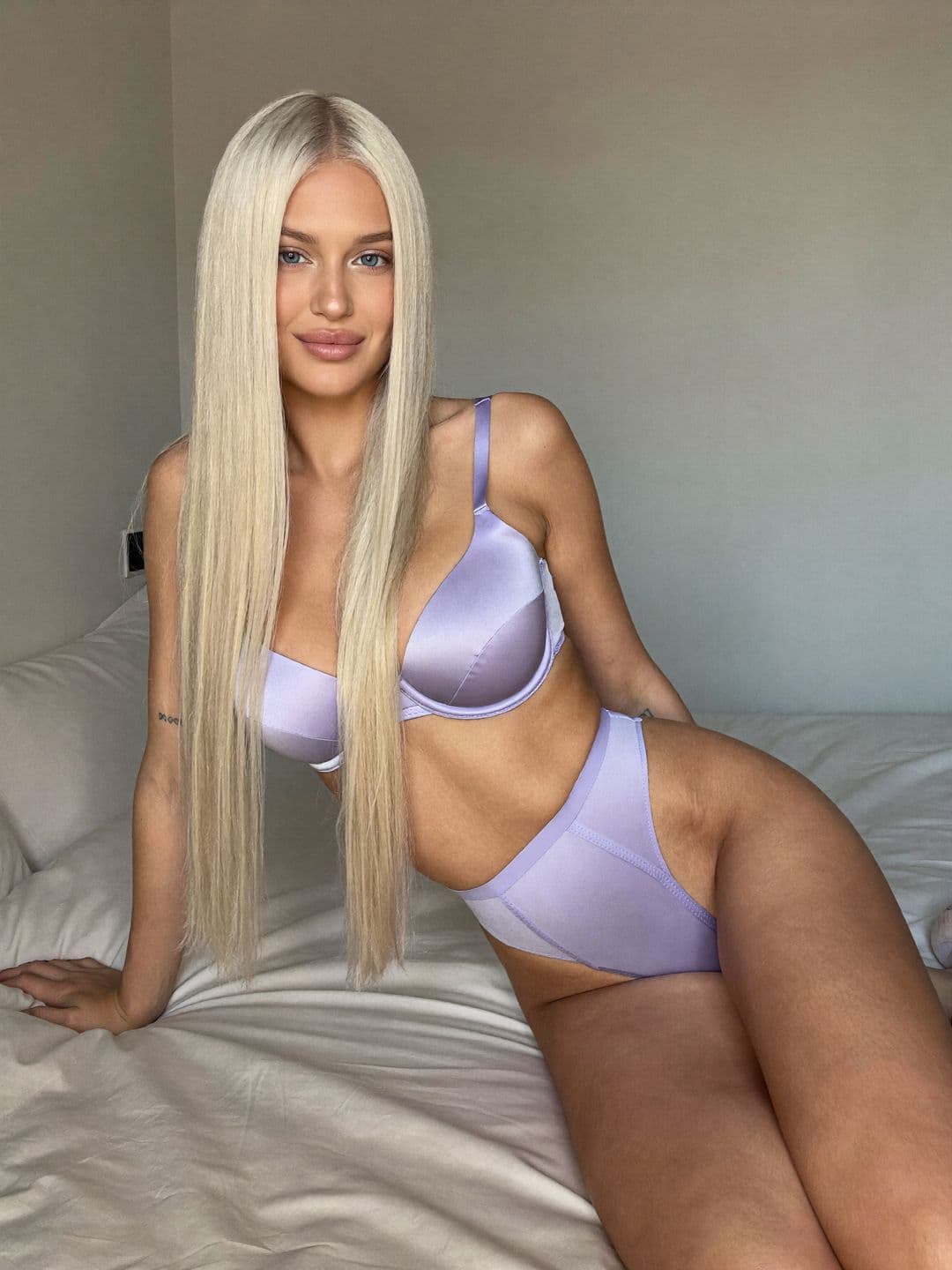 Sofiia, a blonde Ukrainian woman, in lavender satin lingerie lying on her side propped on one elbow across cream sheets