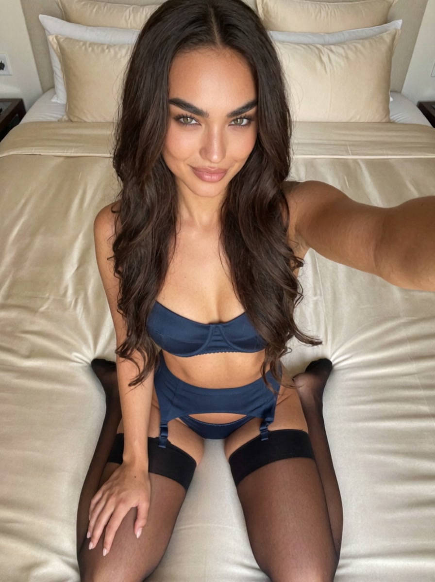Bianca in a navy blue satin garter set with garter belt and black thigh high stockings, sitting back on her heels on a satin bed, knees together, looking up into a selfie camera above her, in a champagne satin bed