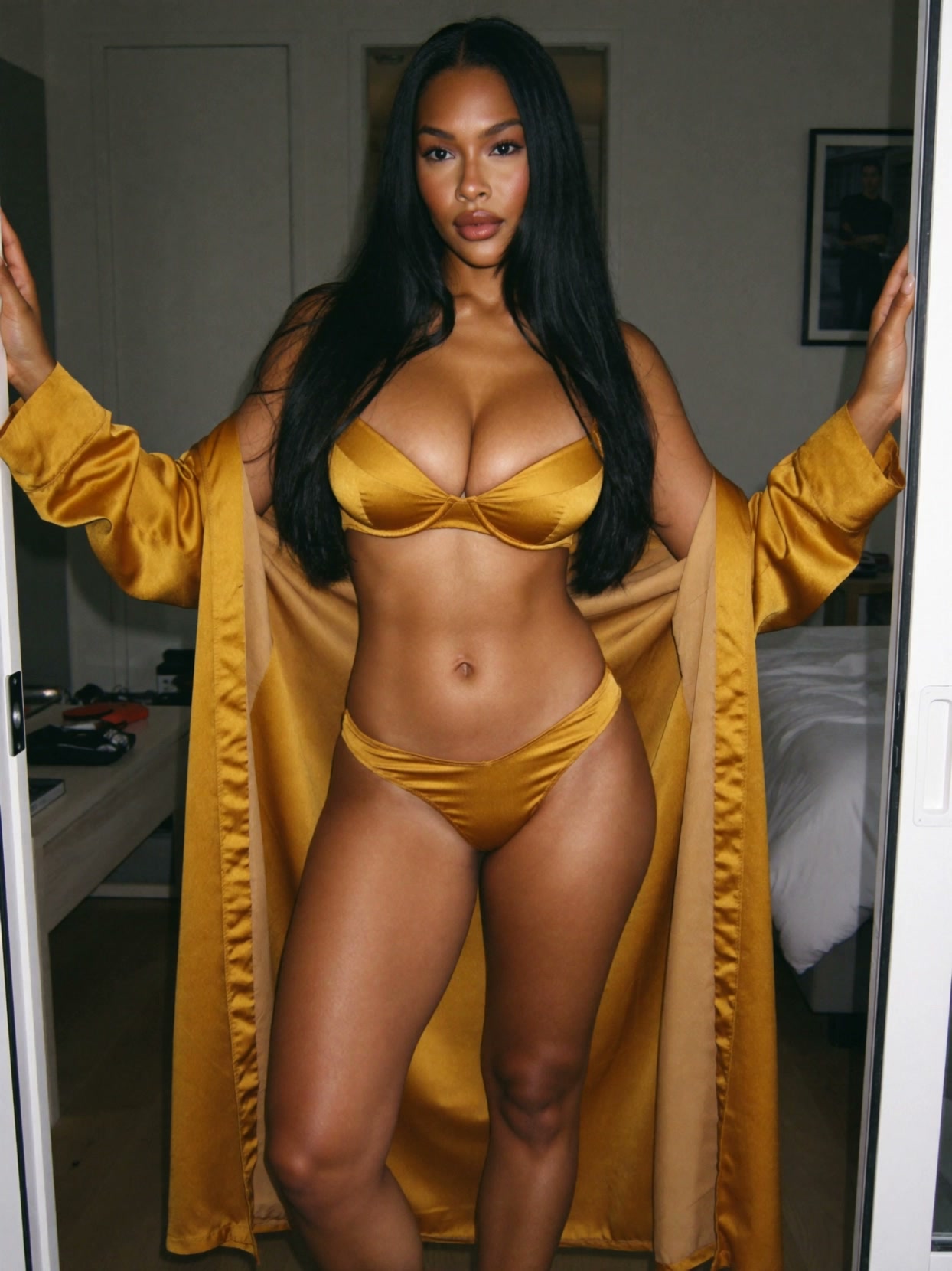 Kira in a gold satin robe open over a matching gold satin bra and briefs, leaning against a doorway with legs together, in a Los Angeles villa bedroom