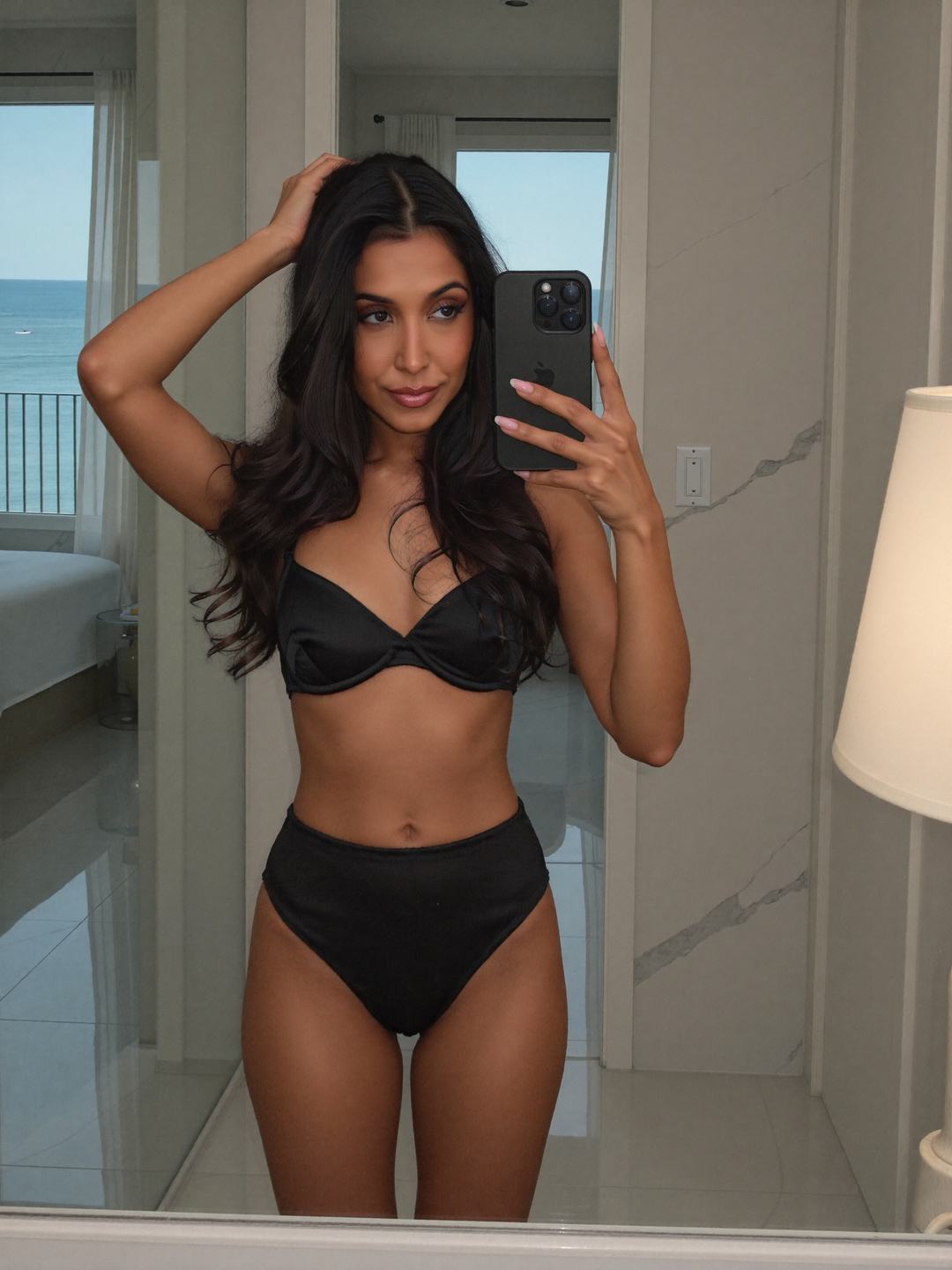 Mirror selfie of Ananya, a dark-haired Indian woman, in a black satin bra and high-waisted black briefs, phone held up in one hand and the other hand on top of her head, marble walls and a balcony with the sea behind her.