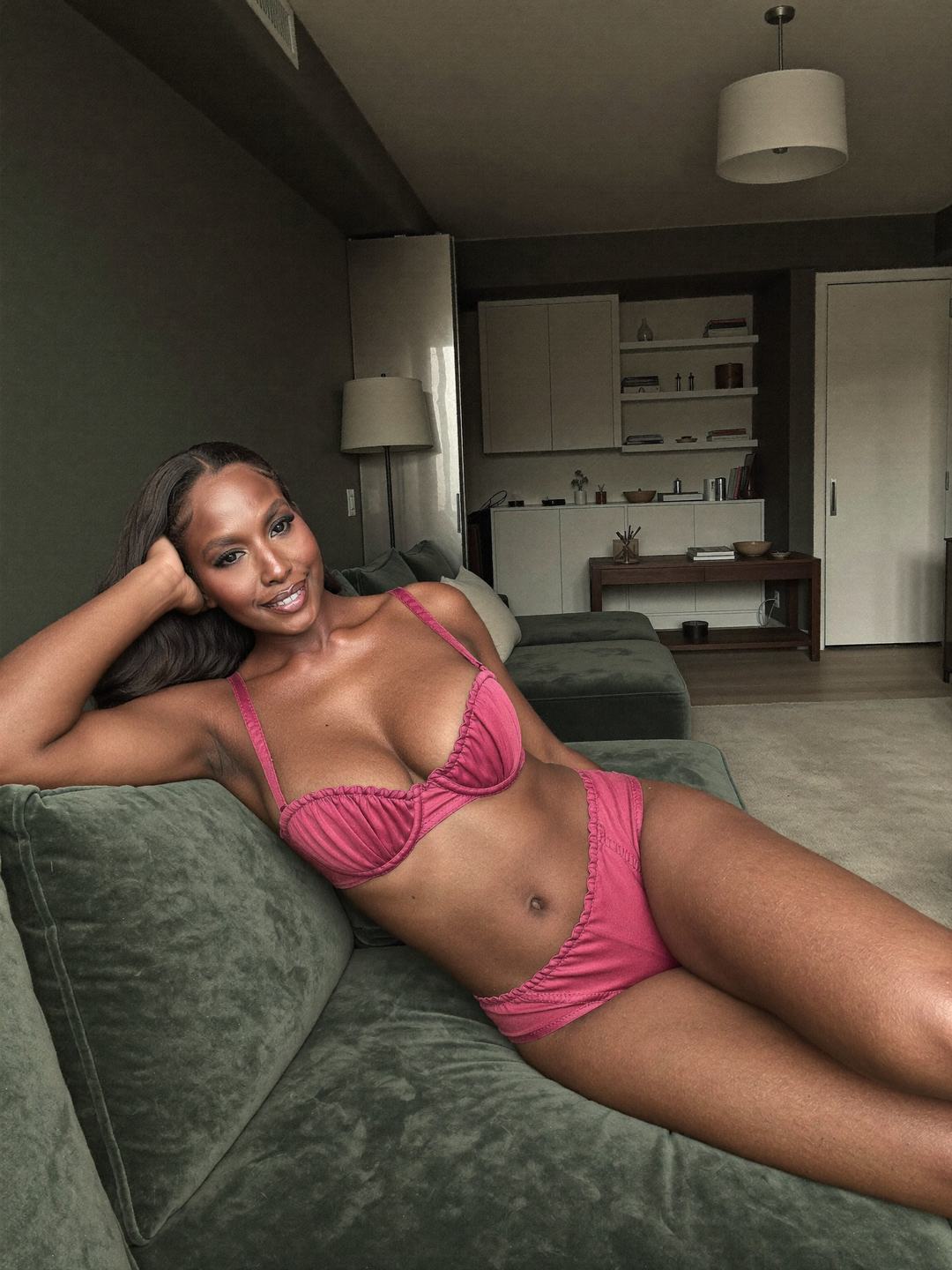 Brielle, a dark-haired African American woman, reclines on a sage green velvet sofa in a rose pink ruched bra and rose pink briefs, one arm behind her head, legs stretched out, a dark green room with shelves behind her, smiling.