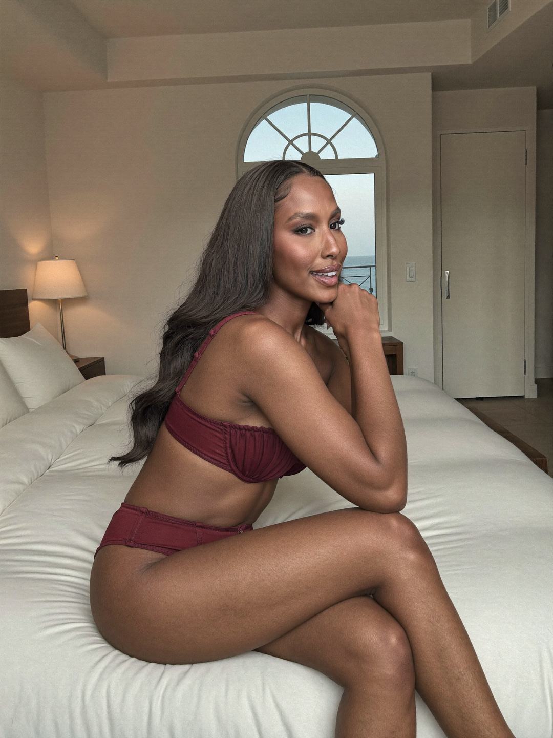 Brielle, a dark-haired African American woman, sits on a white bed in a garnet-red ruched bandeau bra and burgundy briefs, legs crossed, chin on her hand, an arched window and lamp behind her, smiling.