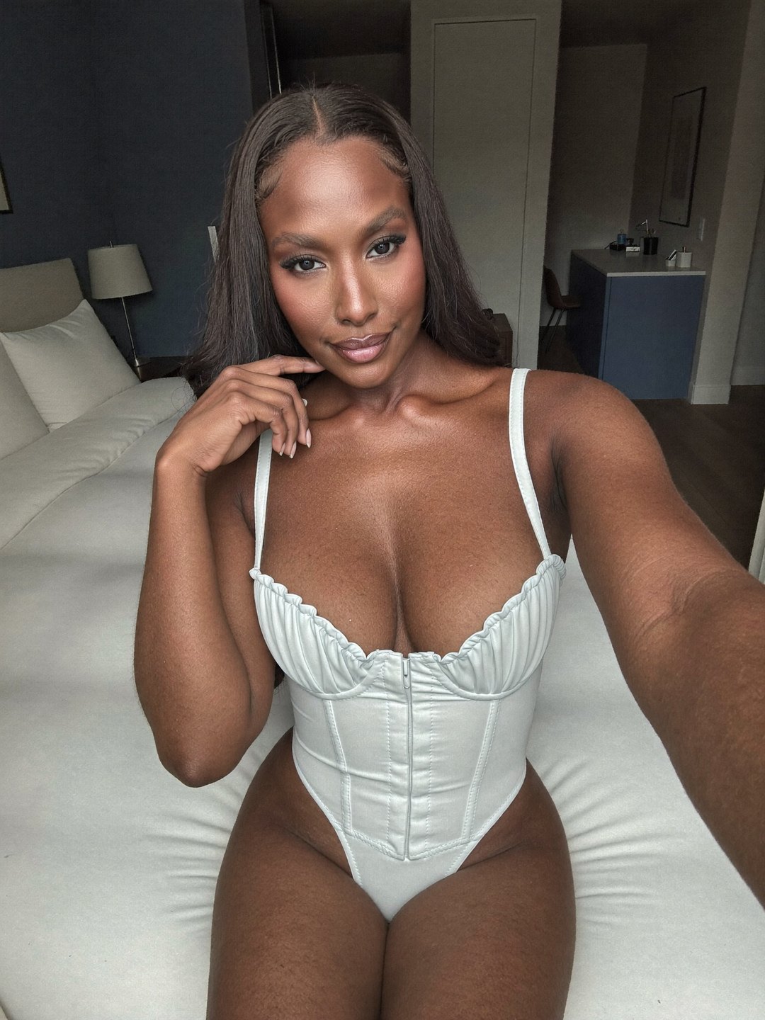 Brielle, a dark-haired African American woman, in an ivory corset bodysuit with ruched cups and a front zip takes an arm's-length selfie seated on a white bed, one hand at her chin, smiling into the lens, a navy wall and blue cabinet behind.