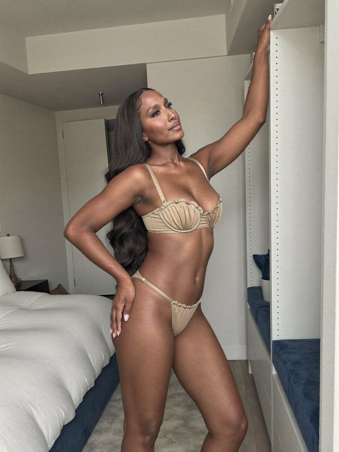Brielle, a dark-haired African American woman, in a sand-beige bra with ruffled shell cups and matching string briefs stands by a white wardrobe, gripping its tall edge overhead, her other palm at her hip, gazing up and away from the camera.