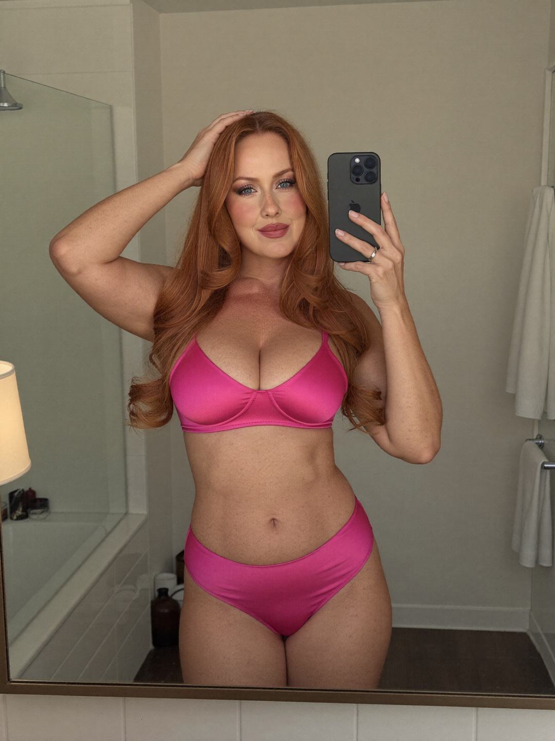 Mirror selfie of Brynn, a redheaded Canadian woman, standing front-on in a hot pink satin bra and hot pink briefs, phone in her right hand, left hand on top of her head, calm smile, a glass shower screen and white towel behind.