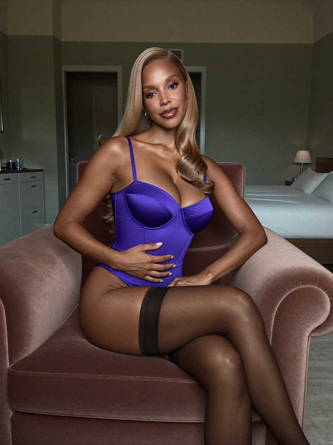 Indigo boned satin corset with matching briefs and black thigh-highs; Danielle, a brunette African American woman, sits in a dusty rose velvet armchair, legs crossed, both hands resting at her waist, smiling, with a white bed behind.