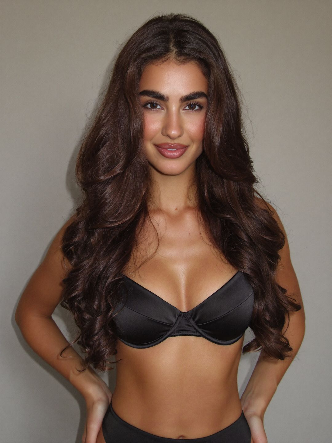Eleni in a black satin bra smiling with her hands on her hips against a warm grey wall