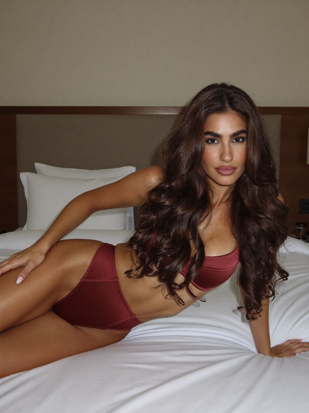Eleni in burgundy satin lingerie lying on her side across a white hotel bed