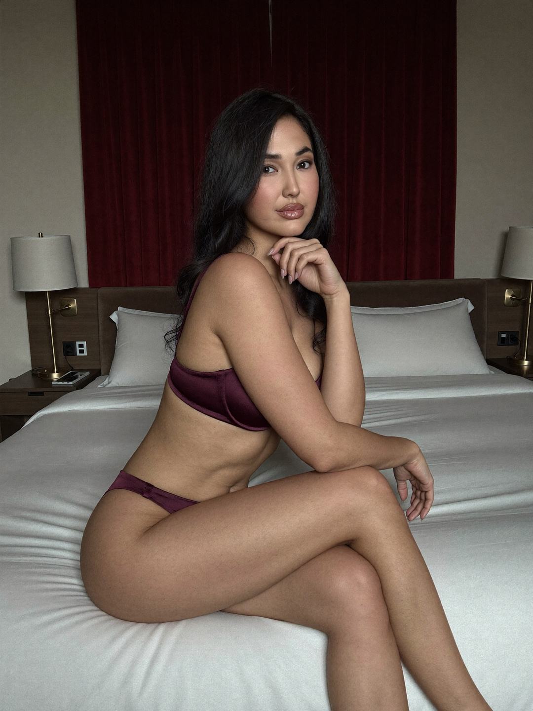 Emi, a dark-haired Japanese woman, sits on the edge of a white bed in a deep plum satin bra and plum briefs, legs crossed, fingers under her chin, calm gaze, red velvet curtains and two wall lamps behind.