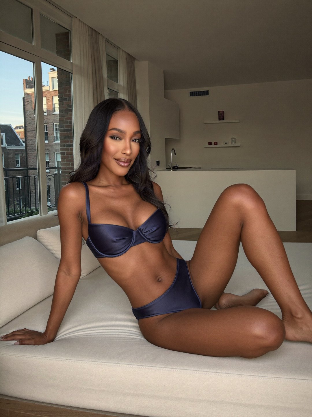 Leaning back on one hand across a deep cream sofa, knees bent, Fabienne, a dark-haired Haitian woman, smiles in an ink blue satin underwire bra and high-cut briefs; a white kitchen and tall curtained windows sit behind.