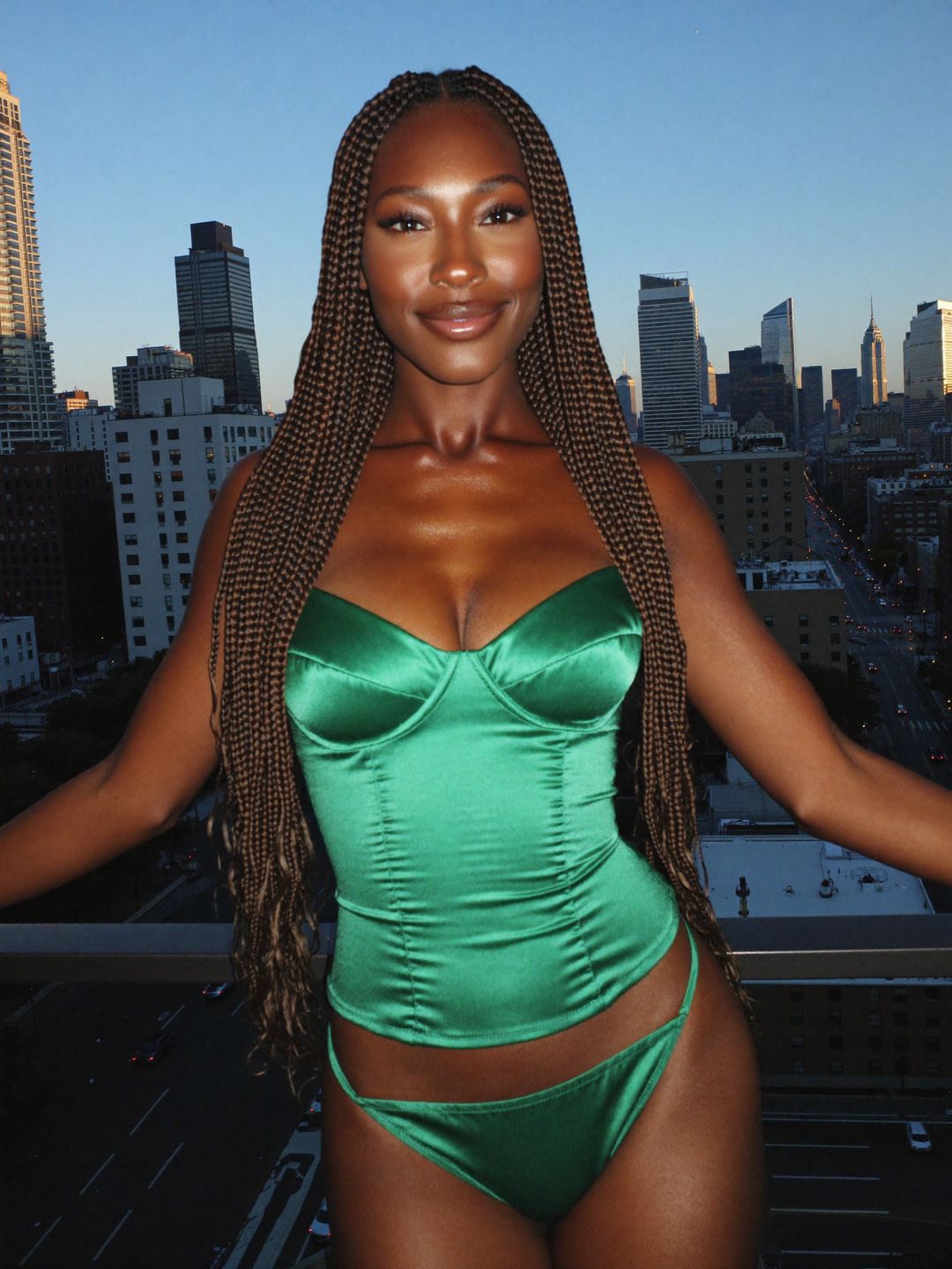 Imani with waist-length dark brown box braids hair, leaning on a railing with a city skyline behind, wearing an emerald satin corset, in a city balcony at blue hour.