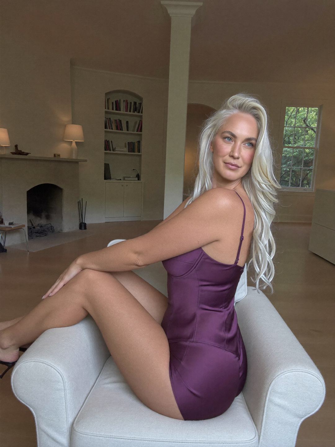 Ingrid, a blonde Norwegian woman, sits sideways on a white armchair in a short purple satin slip, back half turned to the camera, hands on her knee, looking over her shoulder, a fireplace and built-in bookshelves behind.