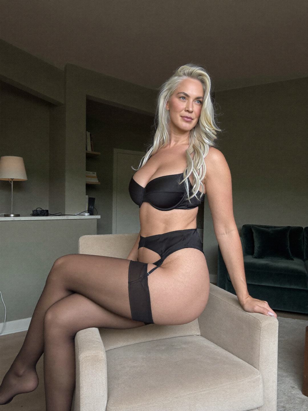 Ingrid, a blonde Norwegian woman, sits on the arm of a beige armchair in a black bra, black garter belt, black briefs and sheer black stockings, legs crossed at the knee, hands on the cushion, a direct look, a dark green sofa behind.