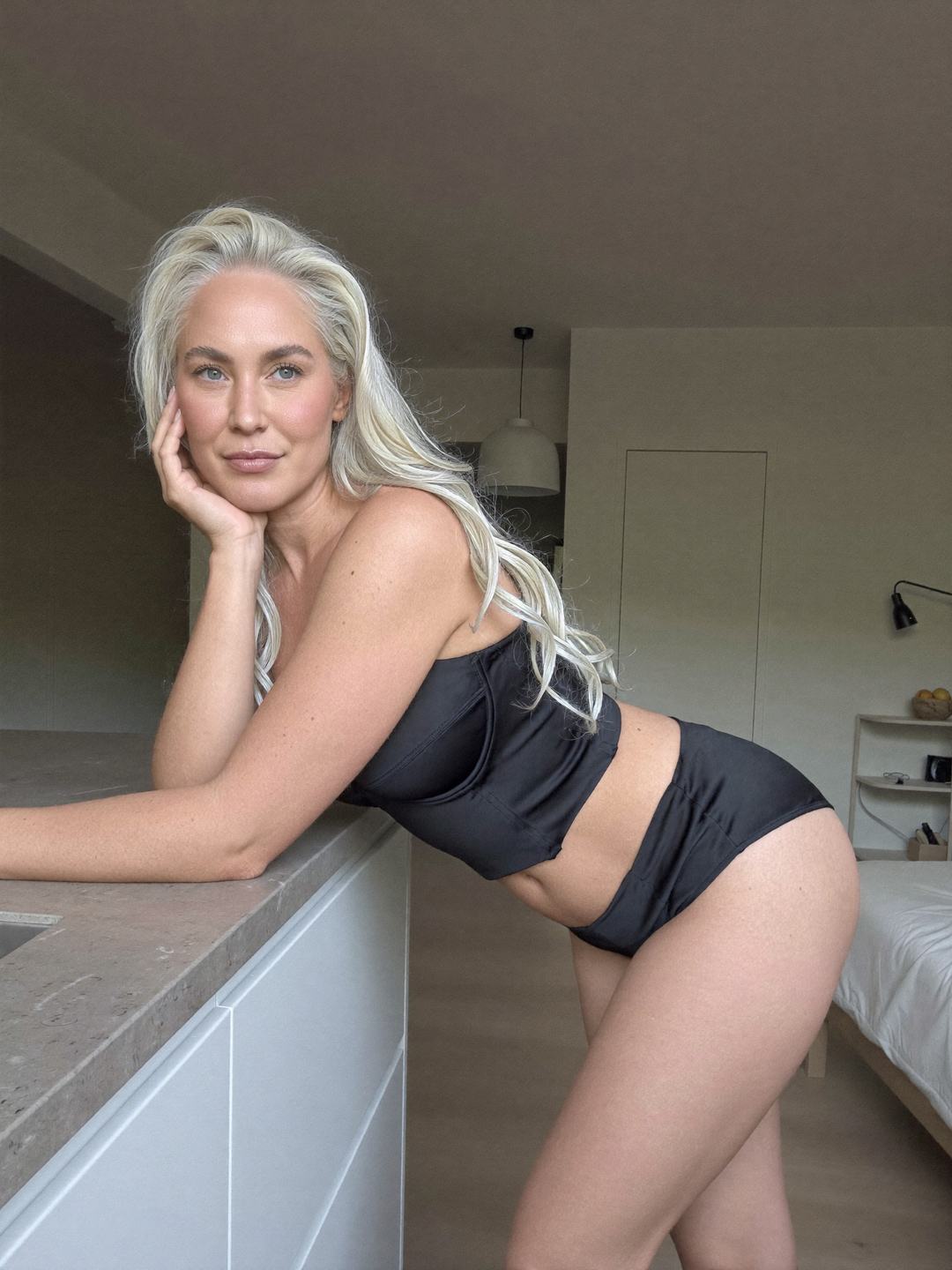 Ingrid, a blonde Norwegian woman, leans forward on a stone kitchen counter in a black satin bandeau top and black high-cut briefs, chin resting on her hand, smiling softly, white cabinets and a bed in the open room behind.