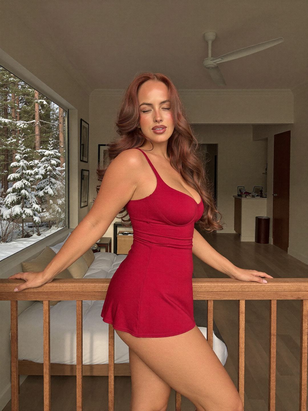 Jacinta, a redheaded Australian woman, stands side-on in a short red slip dress with both hands on a wooden banister, eyes closed with a soft smile, a large window onto snow-covered pine trees and a bedroom with a ceiling fan behind.