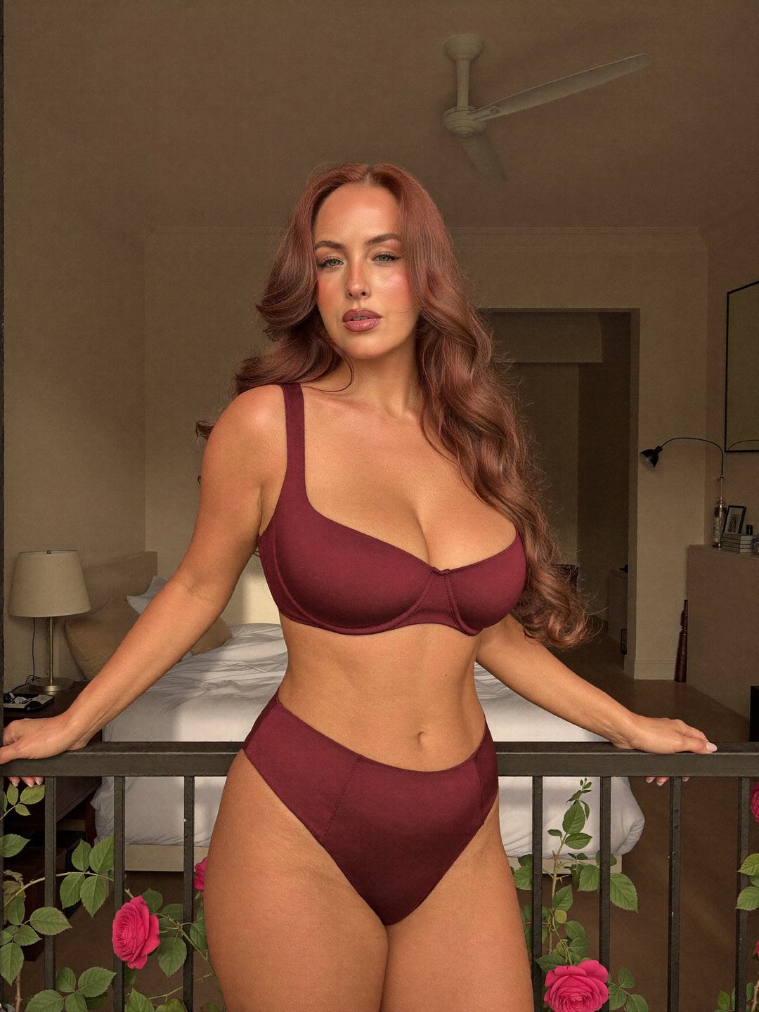Jacinta, a redheaded Australian woman, stands front-on in a burgundy bra and high-waisted burgundy briefs, both hands on a black railing with climbing red roses, steady gaze and closed lips, a bedroom with a lamp and white bed behind her.