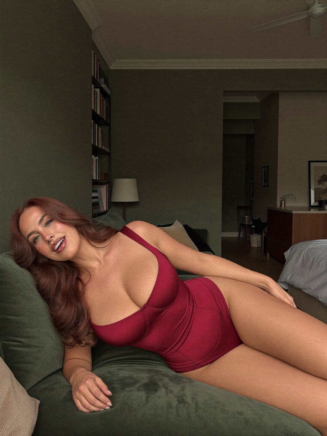 Jacinta, a redheaded Australian woman, reclines on a sage green velvet sofa in a cherry red bodysuit, head tilted back on the cushion, laughing, one hand on her thigh, dark green walls, bookshelves and a lamp behind.