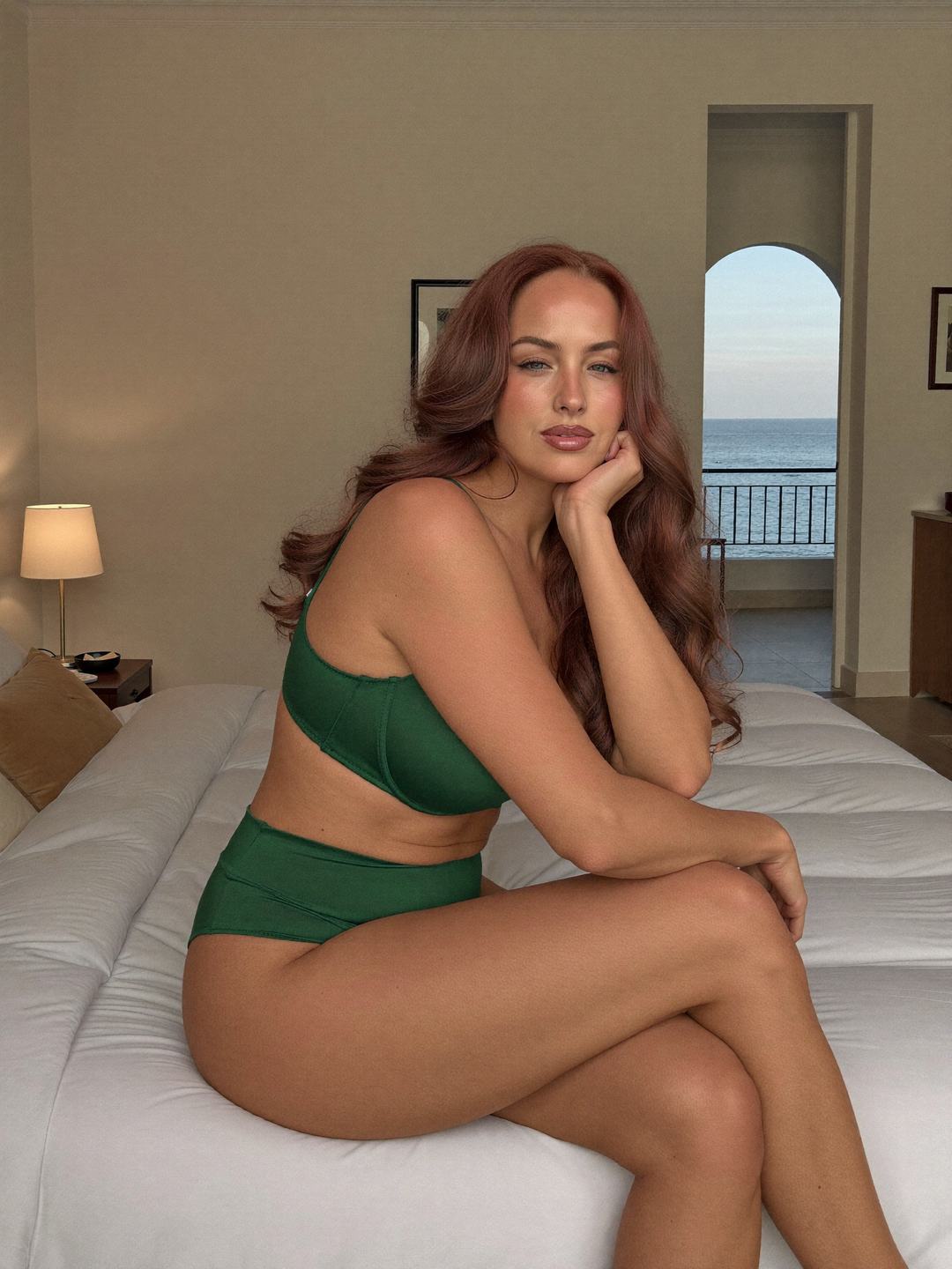 Jacinta, a redheaded Australian woman, sits on the edge of a white bed in a forest green bra and high-waisted green briefs, legs crossed, chin on her hand, soft smile, an arched doorway to a balcony and blue sea behind.