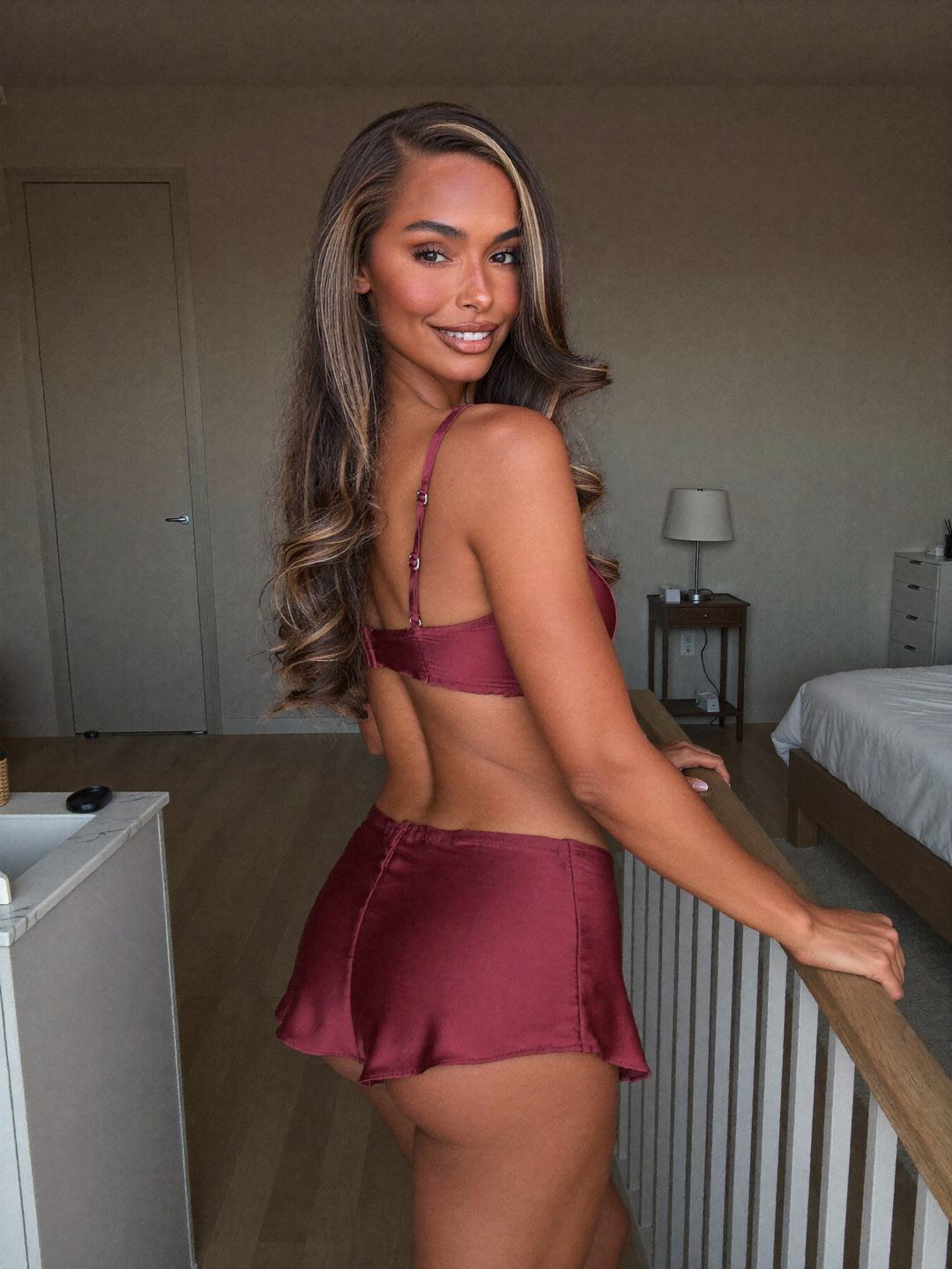 Larissa, a brunette Brazilian woman, standing with her back to the camera beside a white staircase in a burgundy satin bra and flared burgundy satin mini skirt, one hand on the wooden banister, looking back over her shoulder with a smile