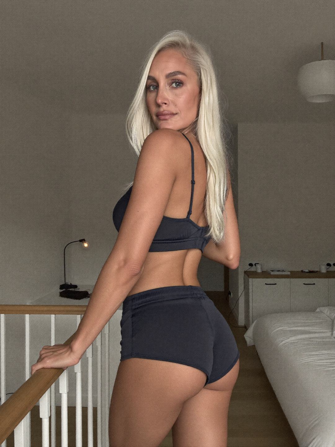 Maren, a blonde Norwegian woman, seen from behind at the top of a white staircase in a charcoal satin bra and charcoal shorts, one hand on the wooden banister, turning her head to look back with a calm expression.