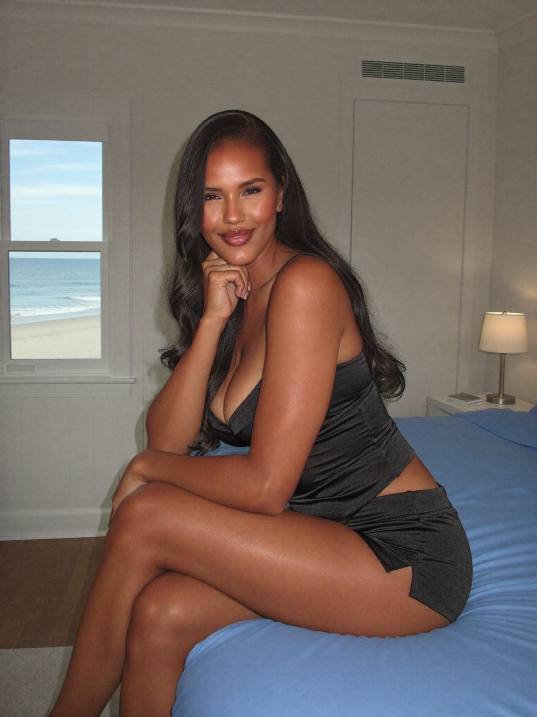 Monique, a dark-haired African American woman, sits on a bed with a sky-blue cover in a black satin camisole and black shorts, legs crossed, hand at her cheek, a window showing sand and ocean, smiling softly.