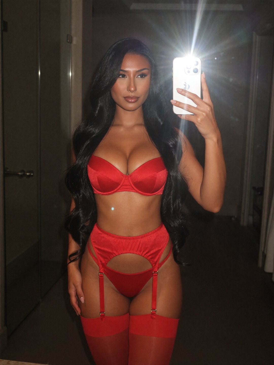 Flash mirror selfie of Reina, a dark-haired Japanese woman, in a red satin balconette bra, red garter belt, briefs and red stockings, phone raised at chest height, hand on her hip, long black hair over her shoulders.