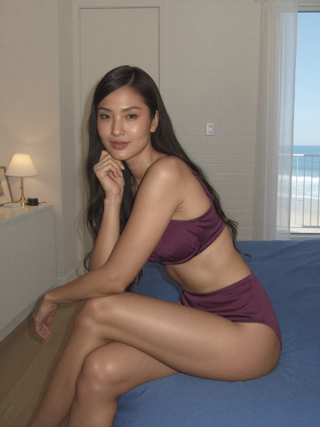 Seoyeon, a dark-haired Korean woman, sits on a bed with a blue sheet in a plum bandeau bra and high-waisted plum briefs, legs crossed, chin on one hand, looking at the camera, an open balcony door with sand and sea behind.
