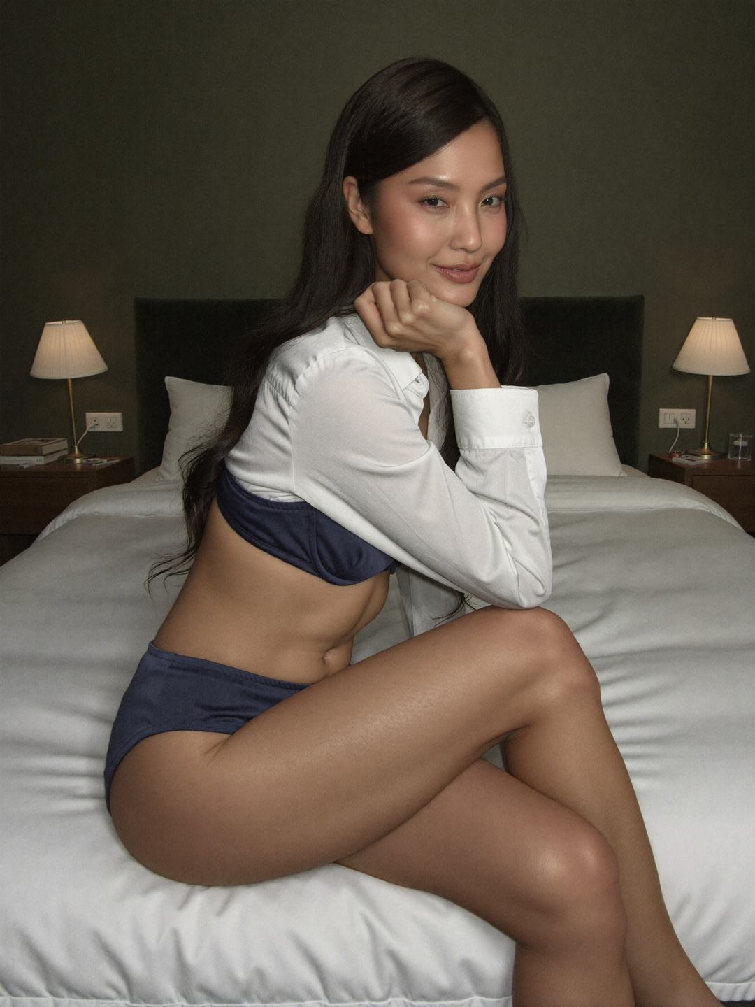 Seoyeon, a dark-haired Korean woman, sits on the edge of a white bed wearing an unbuttoned white shirt over a navy bra and navy briefs, legs crossed, chin on her hand, smiling, two bedside lamps and a dark green wall behind.