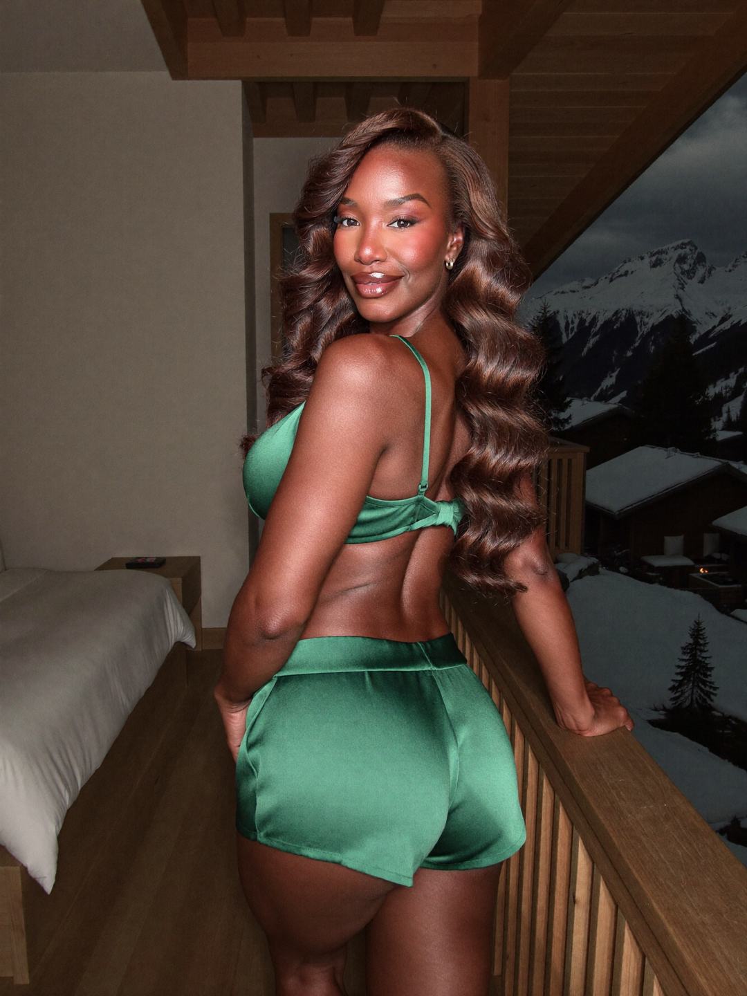 Back view of Simone, a brunette African American woman, in a green satin bra and matching shorts, leaning on a wooden rail indoors, looking back over her shoulder with a smile, snow-covered peaks and chalets beyond the glass.