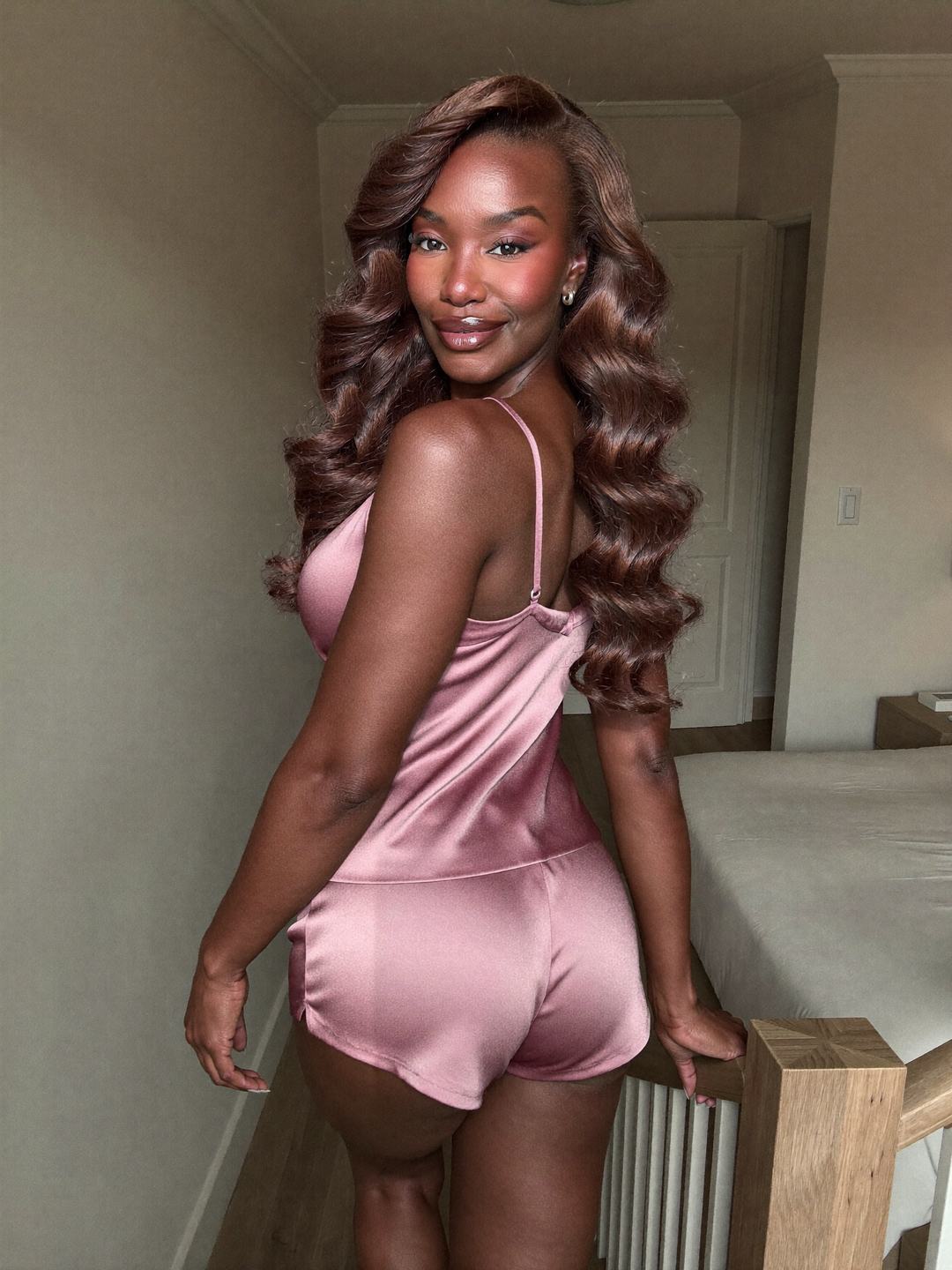Back view of Simone, a brunette African American woman, standing at a wooden stair rail in a pink satin camisole and matching shorts, one hand on the post, turning her head to smile over her shoulder.