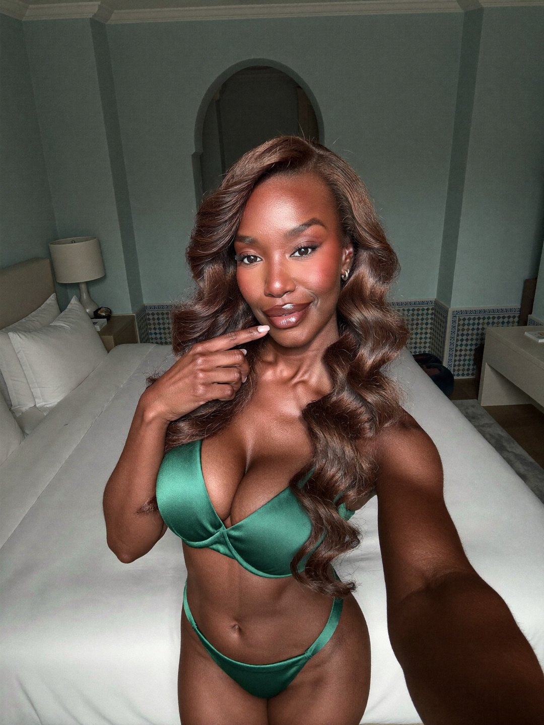 Arm's-length selfie of a woman with long brown waves in an emerald green satin bra and string briefs, finger at her lip, smiling into the phone in front of a white bed and a Moorish arch.