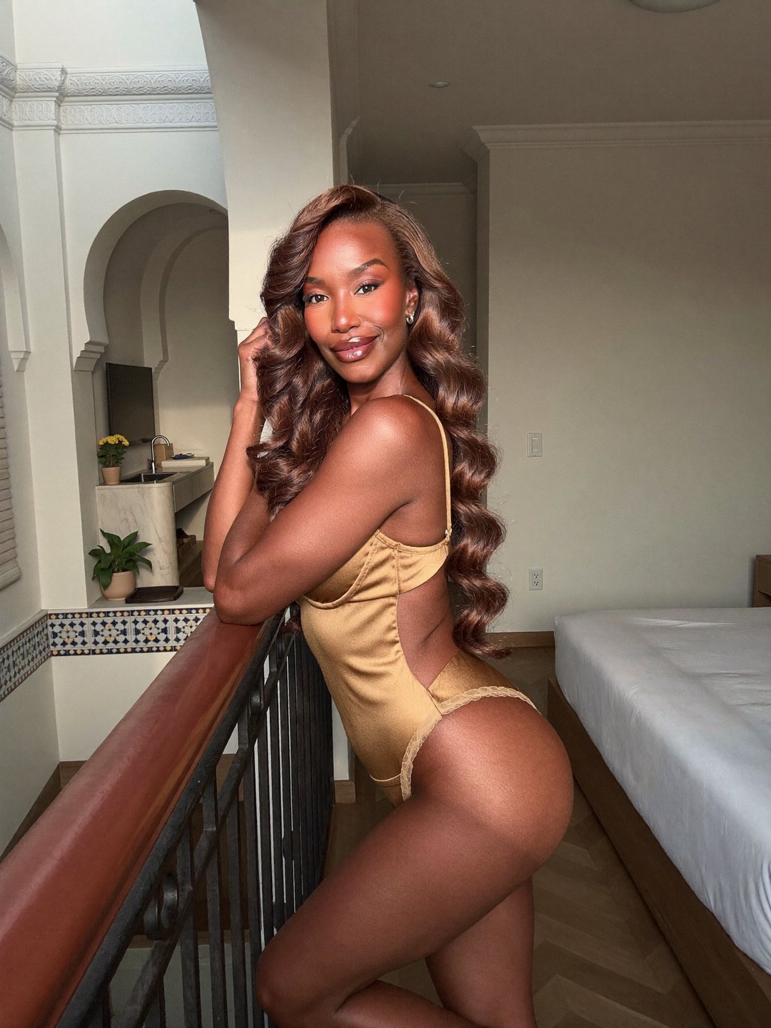 A woman with long glossy brown waves in a gold satin teddy with lace edges leans on a wooden and wrought-iron railing, hand lifted to her hair, smiling over her shoulder, Moorish arches and tiles behind.