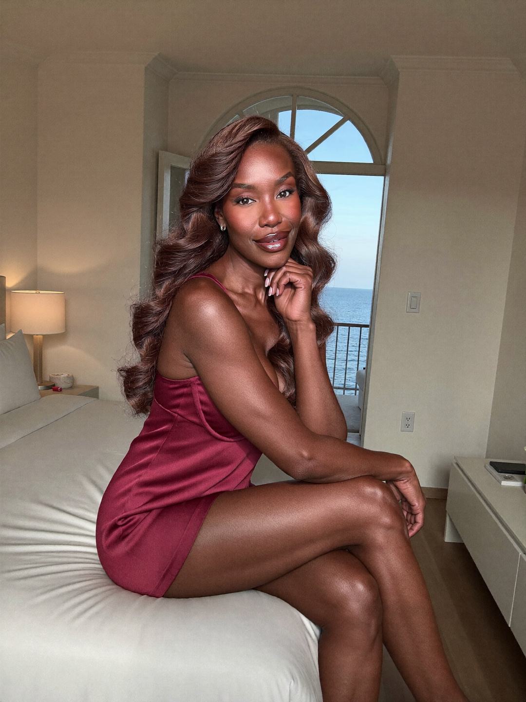 Simone, a brunette African American woman, sits on a white bed in a burgundy satin camisole and shorts, legs crossed, chin on her hand, a tall arched window with the sea beyond, a playful smile.