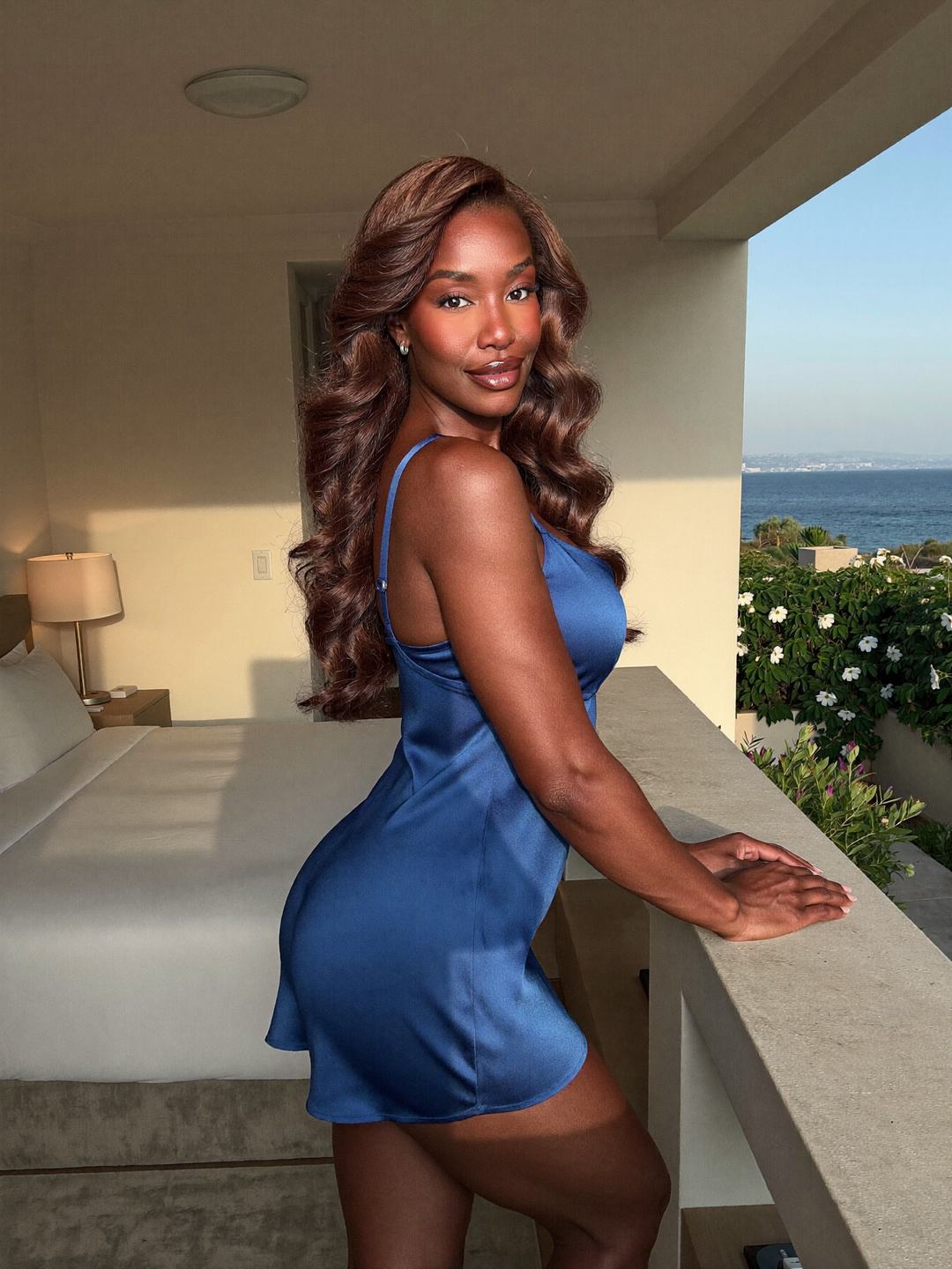 Simone, a brunette African American woman, stands side-on at a low terrace wall in a short cobalt blue satin slip, both hands resting on the ledge, white flowers and the sea behind her, smiling over her shoulder.
