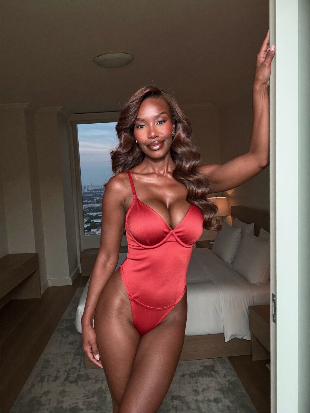 Simone, a brunette African American woman, stands in a doorway in a red satin bodysuit with underwire cups, one arm stretched up along the frame, the other at her side, a bed and a city view behind her, smiling.