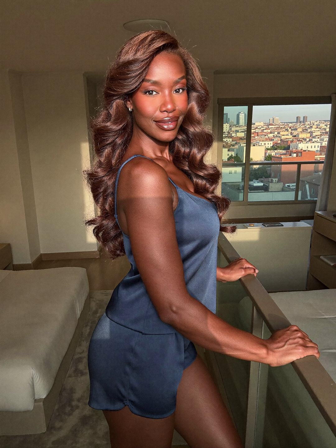 Simone, a brunette African American woman, stands at a balcony rail in a navy satin camisole and shorts, hands on the rail behind her, rooftops of a hillside city in the distance, smiling at the camera.