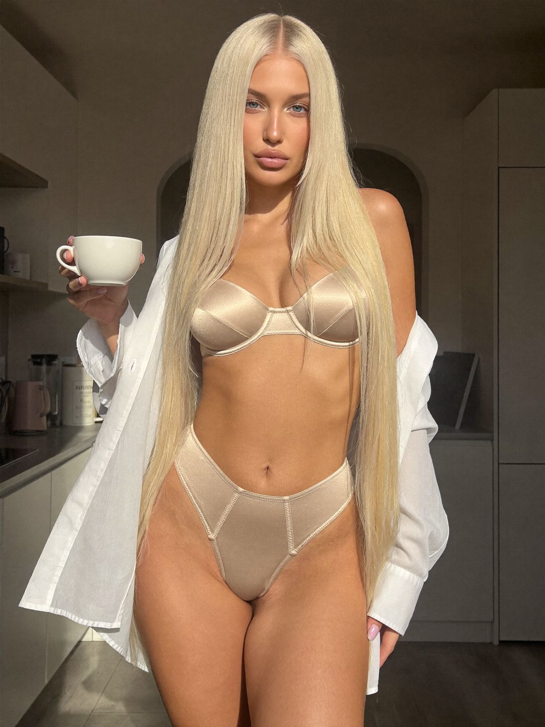 Sofiia in champagne satin lingerie and an open white shirt holding a cup in a sunny kitchen