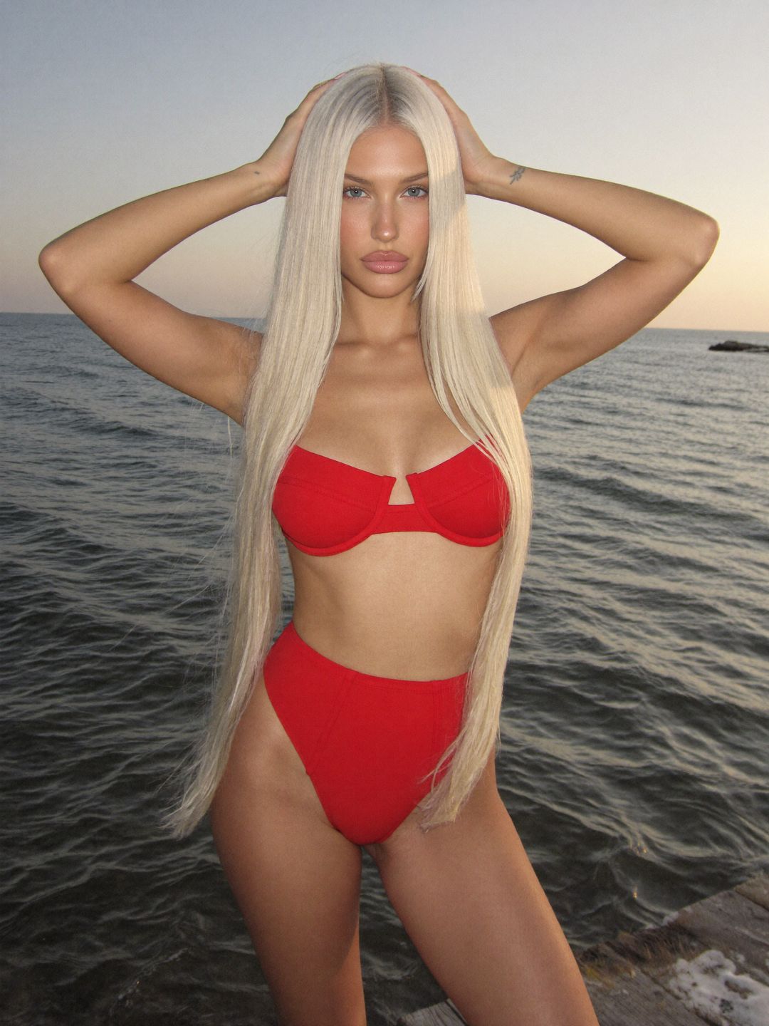 Sofiia in a red bikini with both hands in her hair on a pier over the sea at sunset